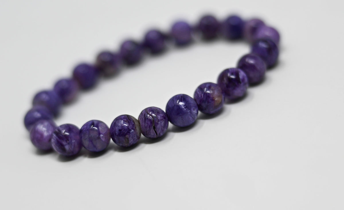 Charoite bracelet with round 8 mm purple beads and pale, mottled and dark vein markings, photographed on a neutral light surface.