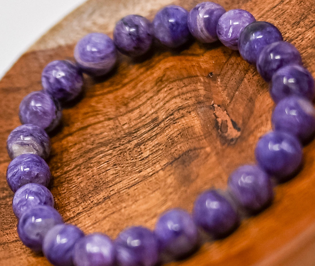 Charoite bracelet with round 8 mm purple beads and varied pale markings, resting on a wooden dish.